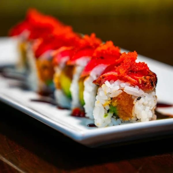 Close-up of a sushi roll with red topping and orange roe on a white rectangular plate.