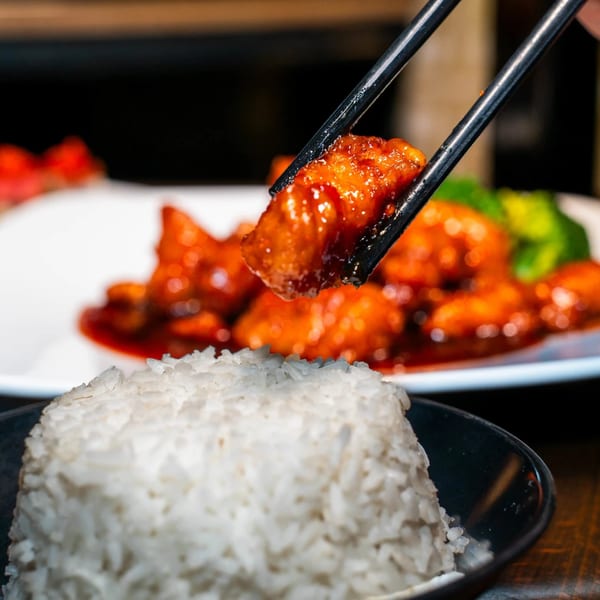 Chopsticks holding a piece of orange chicken over a bowl of white rice at Made in Asia.